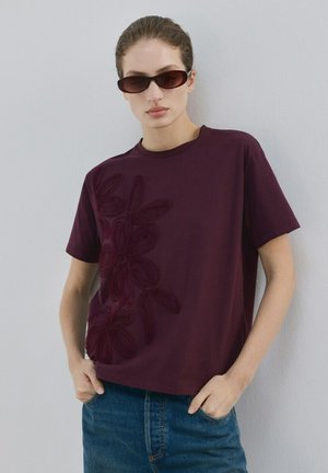 Žena u tamnoplavoj majici s cvjetnim uzorkom i sunčanim naočalama stoji naslonjena na svijetlosivu zid, ruke u džepovima plavih traperica.