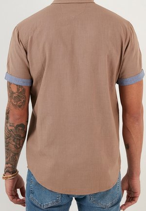 Mężczyzna w beżowej koszuli z krótkim rękawem z niebieskimi podwiniętymi mankietami i niebieskich dżinsach, pokazujący tatuaże na lewej ręce oraz bransoletki na nadgarstku.