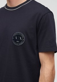 T-shirt noir à manches courtes avec un bord blanc au col et un écusson rond du logo Armani Exchange sur la poitrine.