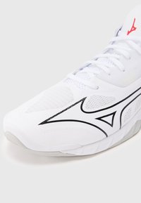 Λευκό αθλητικό παπούτσι με μαύρο γεωμετρικό λογότυπο, τμήματα από δίχτυ, λευκά κορδόνια και κόκκινο έμβλημα του brand στη γλώσσα, εμφανιζόμενο από πλάγια γωνία.