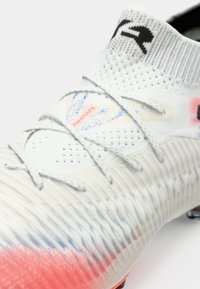 Close-up van een witte voetbalschoen met een gebreide bovenkant, grijze veters, gestructureerde zool en een rood-blauwe verloop bij de teentip.