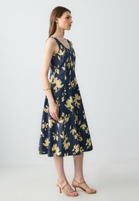 Navyblaues Blumenkleid mit gelben Blumen, ärmelloses Design, A-Linien-Silhouette, knielang, kombiniert mit beigen, geschnürten High Heels.