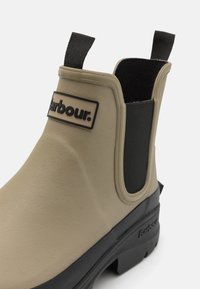 Beige rubber Chelsea boot with a black elastic side panel, textured sole, and a prominent black logo patch on the outer side.