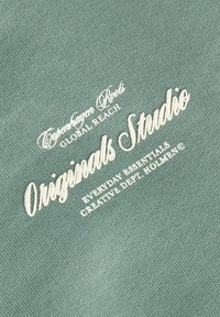 Texte sur tissu vert clair indiquant Copenhagen Roots Global Reach Originals Studio Everyday Essentials Creative Dept Holmen en lettres blanches.