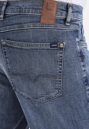 Denim-Jeans in mittlerem Blau mit subtiler, ausgewaschener Textur. Enthält ein hellbraunes Lederetikett am Bund, ein markantes Design der hinteren Taschen und Branding-Details.