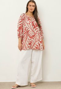 Fließende rote und cremefarbene Tunika mit floralem Muster, ausgestattet mit einem runden Halsausschnitt und einer Bindeschlaufe. Kombiniert mit lockeren weißen Leinenhosen und Sandalen.