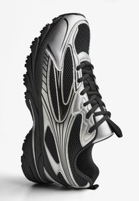 Srebrna i crna sportska cipela s mrežastim panelima, teksturiranim potplatima i izraženim zakrivljenim linijama. Ima crne vezice i ojačani vrh.