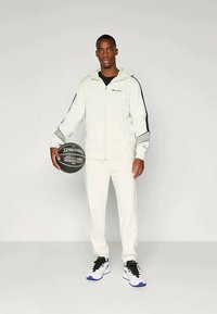 Chándal deportivo blanco con detalles en negro, que incluye una chaqueta con cremallera y pantalones a juego. Sostiene un balón de baloncesto negro y lleva zapatillas negras y blancas.