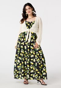 Une femme aux longs cheveux noirs porte une robe maxi noire à motifs de citrons, un cardigan blanc et des talons noirs, posant avec confiance.