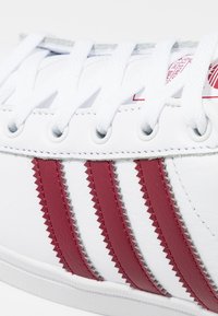 Vita lädersneakers med röda tandade ränder, vita snören och silverfärgade öglor. Har texturerade detaljer längs ränderna.