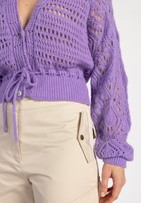Paarse gebreide trui met een ruitpatroon, een taillering en knoopdetails; gecombineerd met beige, high-waisted broek met een klepzak.