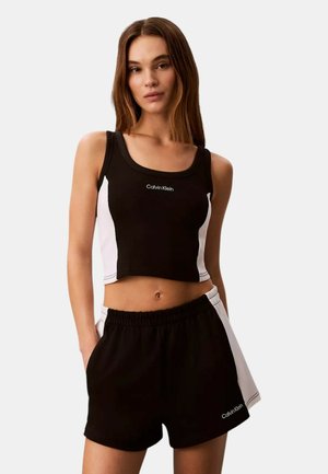 Mujer con top corto sin mangas de Calvin Klein en blanco y negro y pantalones cortos a juego con las manos en los bolsillos contra un fondo liso.