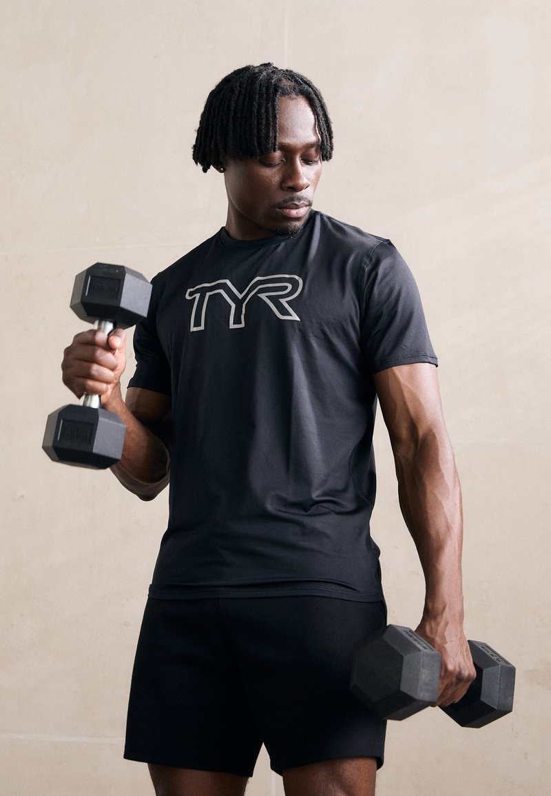Camisa atlética negra con el logo de TYR, combinada con pantalones cortos negros. La persona sostiene mancuernas hexagonales negras, mostrando brazos musculosos.