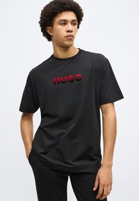 Čierne tričko s krátkym rukávom so výrazným červeným logom "HUGO" v textúrovanom prevedení, vyrobené z hladkej látky s voľným strihom.