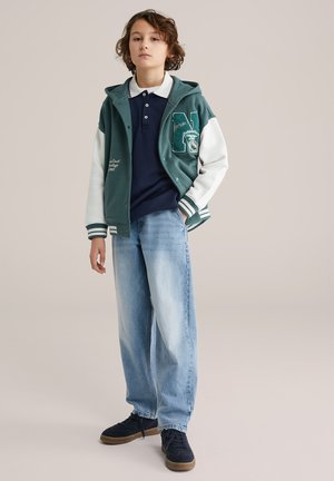 Niño con cabello rizado que lleva una chaqueta varsity verde y blanca sobre un polo azul marino, jeans azul claro y zapatillas negras, de pie con una mano en el bolsillo.