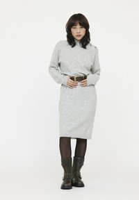 Graues Strickkleid mit hohem Kragen, langen Ärmeln und Knopfdetails an der Schulter. Kombiniert mit einem braunen Gürtel und kniehohen schwarzen Stiefeln.
