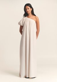Abito lungo monospalla in tessuto beige chiaro con scollatura arricciata e silhouette fluida. Presenta una manica a sbuffo su un lato.