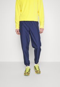 Granatowe spodnie wiatroszczelne z elastycznymi mankietami, połączone z jaskrawożółtym swetrem. Buty są w kolorze beżowym, czarnym i zielonym z neonowymi akcentami.