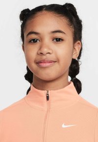 Młodzieżowa kurtka Nike zapinana na zamek, wykonana z miękkiego materiału w kolorze brzoskwiniowym. Posiada wysoki kołnierz, półzamek oraz białe logo na piersi.