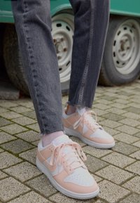 Person wearing light pink and white Nike sneakers with pink socks and dark grey jeans standing on a paved surface near a vehicle wheel.