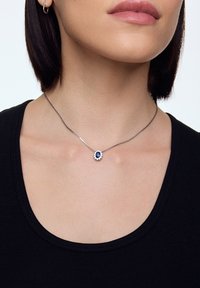 Srebrny naszyjnik łańcuszkowy z owalnym niebieskim kamieniem otoczonym małymi przezroczystymi kamieniami, noszony na czarnej koszuli w zestawieniu z neutralnym odcieniem skóry.