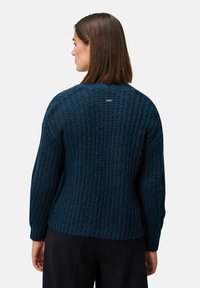 Frau mit glattem braunen Haar, die einen dunkelblauen strukturierten Strickpullover und schwarze Hosen trägt, von hinten vor einem schlichten Hintergrund gezeigt.