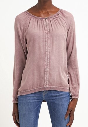 Frau trägt eine langärmlige Bluse in staubigem Rosa mit vorderer Faltenlegung und elastischem Ausschnitt, kombiniert mit blauen Jeans.