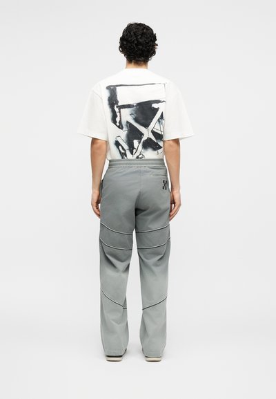 Άνδρας που φοράει άσπρο μπλουζάκι με μαύρο αφηρημένο σχέδιο στην πλάτη και φαρδιά γκρι παντελόνια με ραμμένες λεπτομέρειες, στέκεται γυρισμένος με την πλάτη.