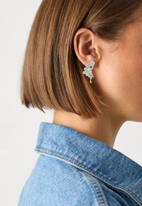 Orecchini floreali argentati con molteplici piccoli embellishments e un accessorio pendente, indossati all'orecchio di una donna, con una giacca di jeans sullo sfondo.