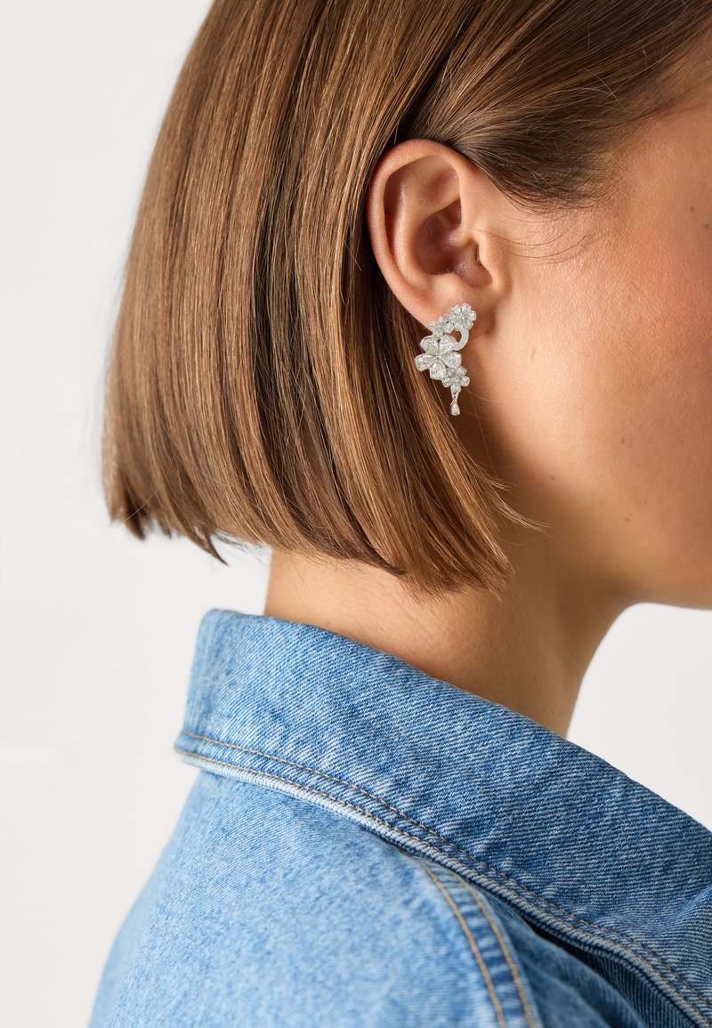 Orecchini floreali argentati con molteplici piccoli embellishments e un accessorio pendente, indossati all'orecchio di una donna, con una giacca di jeans sullo sfondo.