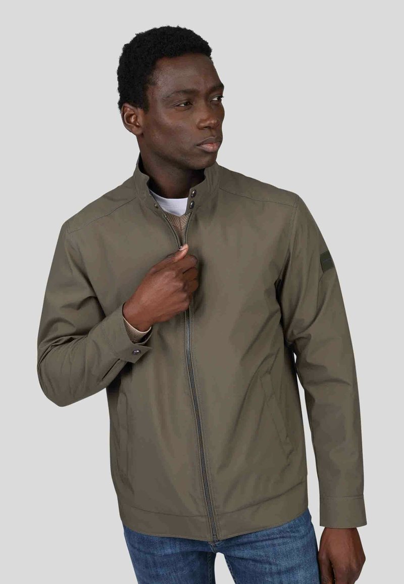 Veste zippée vert olive avec col montant, poignets côtelés et poches latérales. Tissu lisse et léger avec des détails de couture subtils.