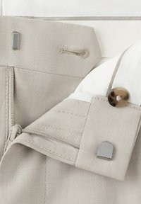 Vaaleanbeiget housut, joissa on vyötärönauhan hakasuljin, vetoketju, nappi ja kankainen lenkki yksityiskohtana, valkoisella pinnalla.