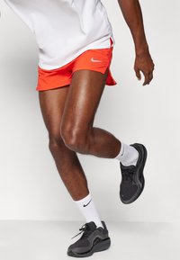 Røde sportsshortser med sidespalter, kombinert med svarte sneakers og hvite sokker. Nike-logoen er synlig på shortsen og skoene.