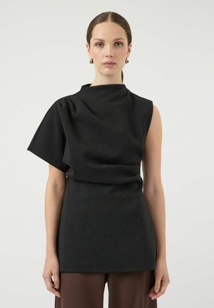 Vrouw met naar achteren gekamd haar, gekleed in een zwarte asymmetrische mouwloze top met één brede mouw, gecombineerd met een bruine broek, tegen een effen achtergrond.