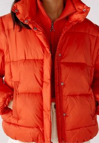 Veste matelassée rouge avec texture matelassée, boutons-pression et col montant. Superposée à un pull zippé assorti avec détails côtelés.