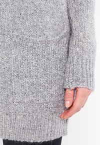 Gros plan sur une main reposant à côté de la poche d'un pull en laine gris avec des poignets et un ourlet côtelés.