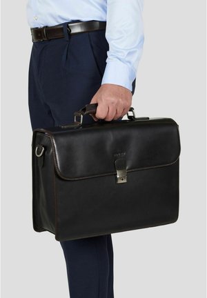 Man in lichtblauw overhemd en donkerblauwe broek houdt een gesloten zwarte leren aktetas bij het handvat vast tegen een grijze achtergrond.