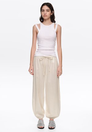 Mujer con cabello largo y oscuro que lleva una camiseta sin mangas blanca, pantalones sueltos color crema con cordón y zapatillas grises abiertas, de pie frente a un fondo liso.