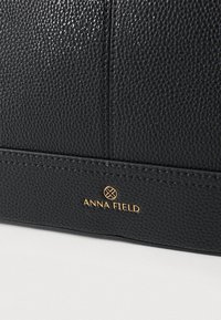 Crna preklopna torba teksture, izrađena od sintetičkog materijala, s vidljivim zlatnim logotipom "ANNA FIELD" i zakrivljenim detaljima šavova.