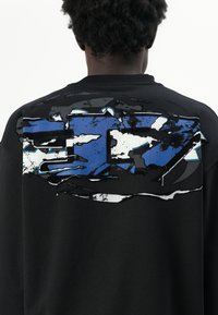 Sweat-shirt noir avec un design abstrait texturé en bleu et blanc au dos. Le col est côtelé et le tissu semble doux.