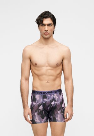 Homme musclé aux cheveux foncés portant un boxer Björn Borg violet et noir à motifs, debout devant un fond clair uni.