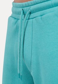 Turkusowe spodnie dresowe mają teksturowany pas i sznurki, gładkie wykończenie tkaniny oraz boczne kieszenie użytkowe.