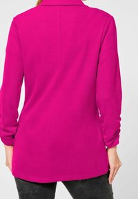Długi rękaw, bluzka bez kołnierzyka w intensywnym fuksjowym kolorze, wykonana z gładkiego dzianinowego materiału o prostym kroju, pokazana od tyłu.