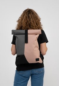 Rullad ryggsäck i grå och rosa tyg, med ett svart bälte och detaljer. Slät yta, rektangulär form med ett rymligt fack.