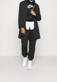 Black overcoat over a black hoodie with white branding, paired with a white shirt, black cargo pants, and white sneakers. Textured handbag detail.