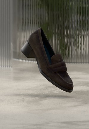 Donkerbruine suède loafers met hak, ronde neus en blokhak tegen een wazige moderne getextureerde achtergrond.