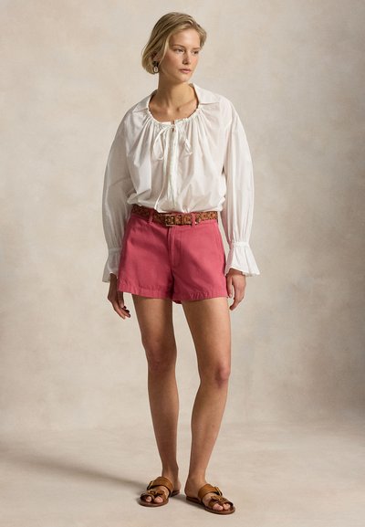 Γυναίκα που φοράει λευκό μπλουζάκι με μακριά μανίκια και βολάν στα μανίκια, ροζ σορτς με πλεκτή καφέ ζώνη και καφέ φλατ σανδάλια, στέκεται μπροστά σε ουδέτερο φόντο.