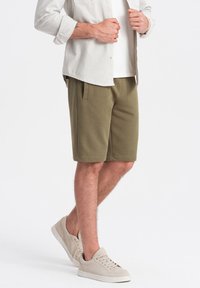 Shorts en mélange de coton vert olive avec poches latérales, taille plate et coupe décontractée, associés à des baskets beiges et une chemise de couleur claire.