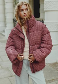 Bordeaux quilted pufferjakke med høj krave, lynlås og trykknapper. Båret over en lys sweater og parret med lyse jeans.