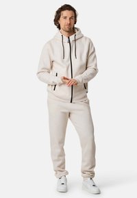 Ljust beige hoodie och matchande joggers i mjukt material, med svart dragkedja, dragsko och sidofickor. Bärs med vita sneakers.